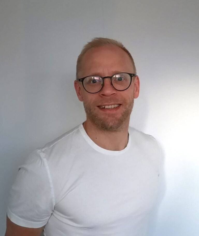 Mann med briller smilende i hvit t-skjorte foran lys bakgrunn.