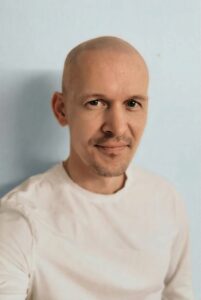 Mann med skallet hode og hvit t-skjorte smilende mot kamera.