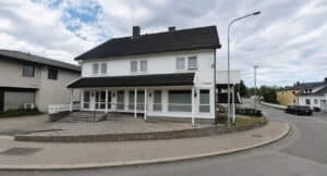 Hvit to-etasjes bolig med veranda og parkeringsplass i forgrunn.