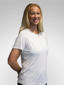 Kvinne smiler i hvit t-skjorte, står mot lys grå bakgrunn.