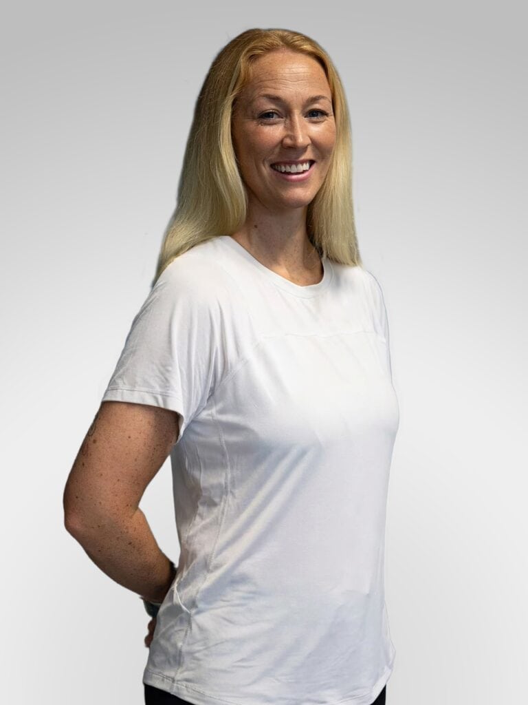 Kvinne smiler i hvit t-skjorte, står mot lys grå bakgrunn.