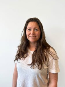 Kvinne med langt brunt hår smiler mot kamera, hvit bakgrunn.