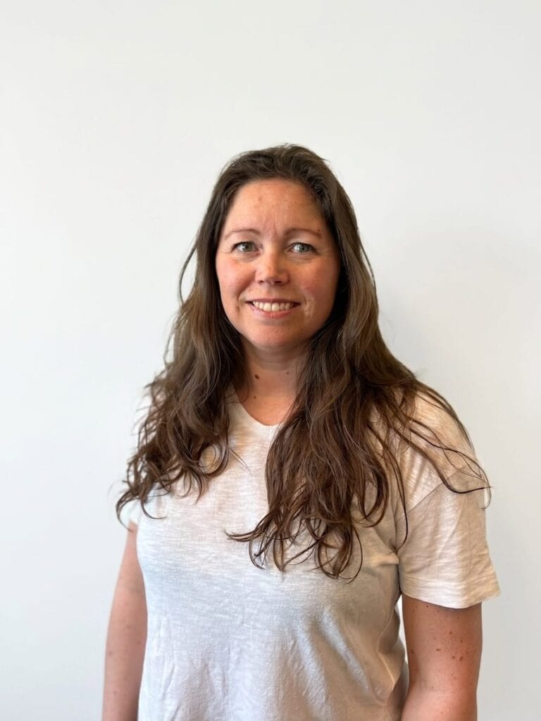 Kvinne med langt brunt hår smiler mot kamera, hvit bakgrunn.
