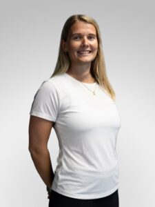 Kvinne smilende i hvit t-skjorte, portrett mot lys grå bakgrunn.