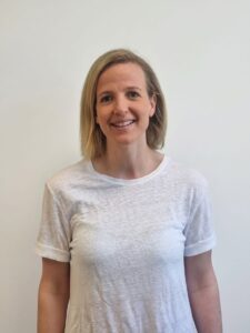 Kvinne smiler i hvit t-skjorte mot en lys vegg.
