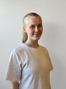 Kvinne med blondt hår i hvit t-skjorte smiler mot kamera.