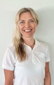 Smilende kvinne med blondt hår i hvit polo-shirt mot lys bakgrunn.