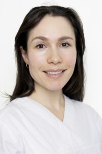 Kvinne i hvit klinikkuniform smiler mot kamera.