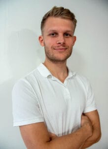 Ung mann i hvit polo smilende med armene krysset mot hvit vegg.