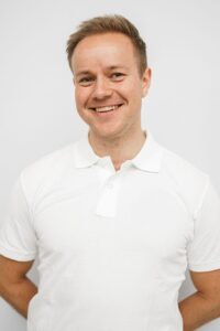 Mann med hvit polo smiler mot kamera, portrettbilde.