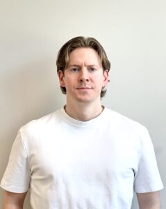 Mann i hvit t-skjorte ser direkte inn i kamera mot en lys bakgrunn.