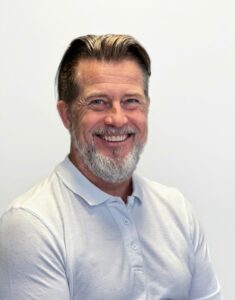 Profesjonell mann med grått hår og skjegg smiler mot kamera.