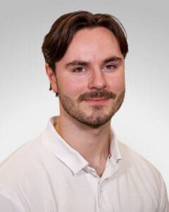 Ung mann med mørkt hår og skjegg i hvit skjorte smilende mot kamera.