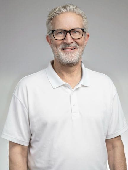 Mann med grått hår og skjegg i hvit polo, smilende, mot lys grå bakgrunn.