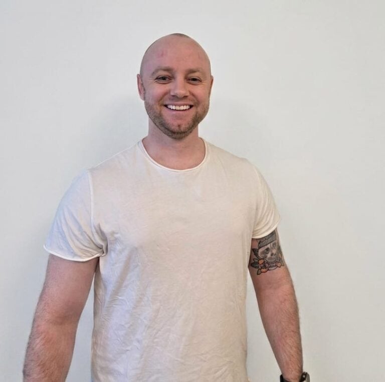 Mann med tatovering smiler i hvit t-skjorte mot hvit bakgrunn.