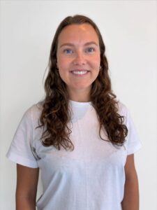 Kvinne med langt brunt hår smilende mot kamera, hvit bakgrunn.
