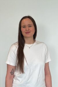 Kvinne med lang brunt hår i hvit t-skjorte smilende mot kamera.