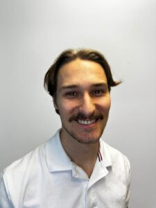 Ung mann med mørkt hår og skjegg smiler mot kamera, hvit bakgrunn.