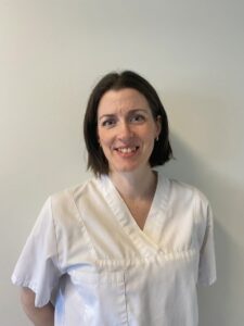 Kiropraktor smiler i hvit klinikkuniform mot en lys vegg.