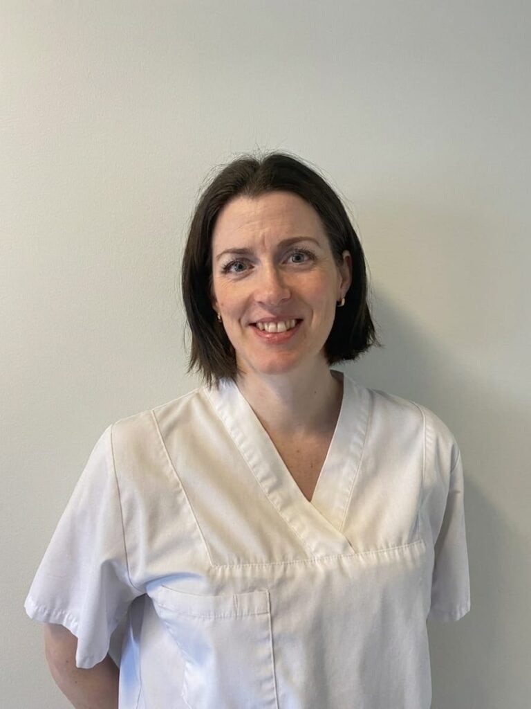 Kiropraktor smiler i hvit klinikkuniform mot en lys vegg.