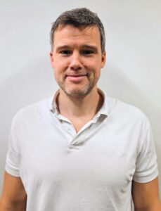 Mann med kort hår og hvit polo shirt smilende mot kamera.