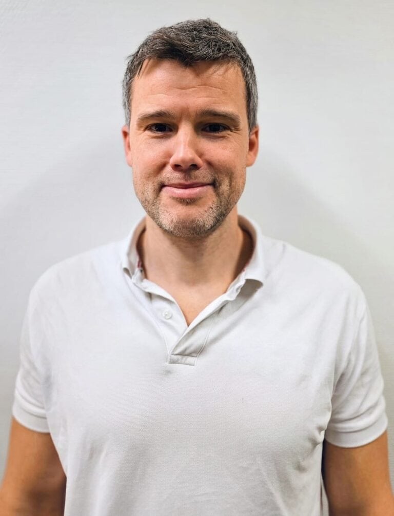 Mann med kort hår og hvit polo shirt smilende mot kamera.