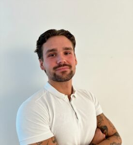 Mann med tatoveringer i hvit polo, smilende, mot en lys bakgrunn.
