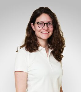 Kvinne med briller smilende i hvit skjorte mot lys bakgrunn.