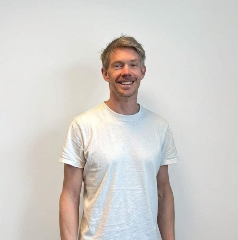 Mann smiler i hvit t-skjorte foran en hvit vegg.