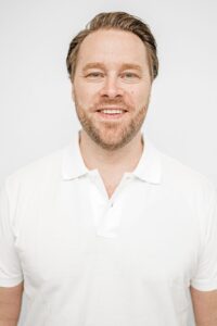 Mann med lyst hår og skjegg smilende i hvit polo.