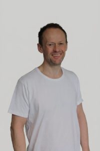 Mann smiler i hvit t-skjorte mot hvit bakgrunn.