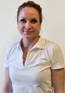 Kvinne med hvit polo smilende mot kamera, innendørs.