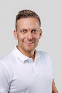 Mann med kort hår i hvit polo smiler mot kamera.