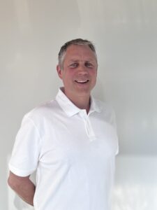 Kvinne i hvit polo-shirt smiler, står innendørs.