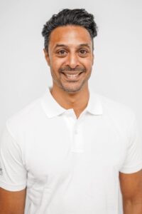 Mann med mørkt hår og hvit polo-shirt smilende mot kamera.