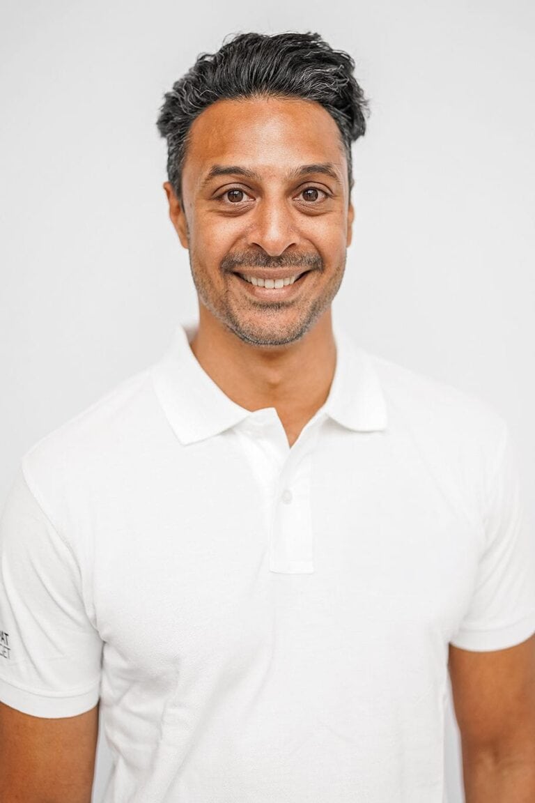 Mann med mørkt hår og hvit polo-shirt smilende mot kamera.