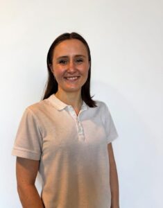 Kvinne smilende terapeut i hvit polo foran hvit vegg.