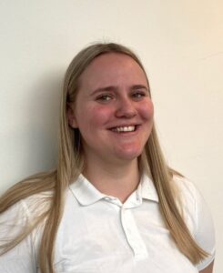 Smilende kvinne med langt blondt hår i hvit skjorte mot en lys vegg.