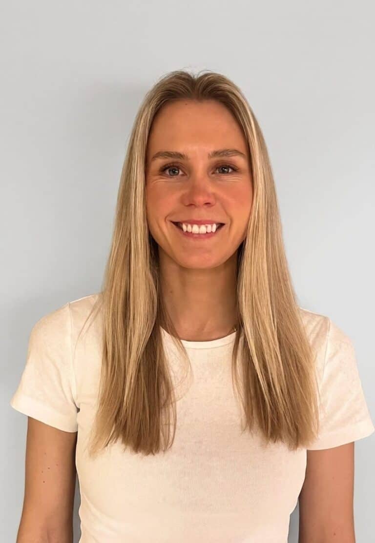 Smilende kvinne med langt blondt hår i hvit t-skjorte foran lys bakgrunn.