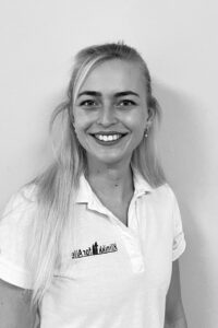 Kvinne smiler til kamera, iført hvit arbeidsuniform, i klinikkmiljø.