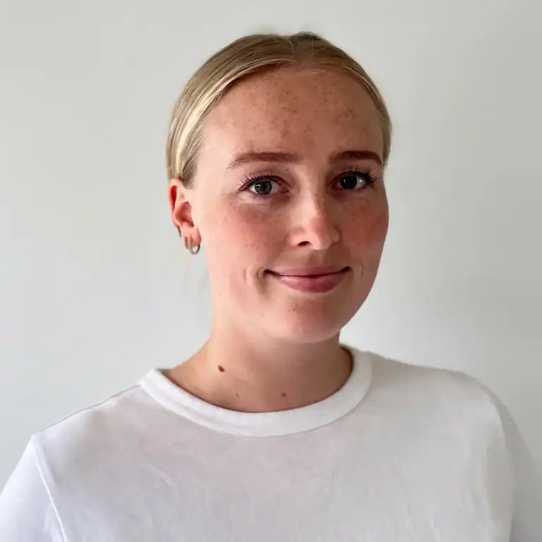 Ung kvinne med blondt hår og hvit t-skjorte smiler mot kamera.